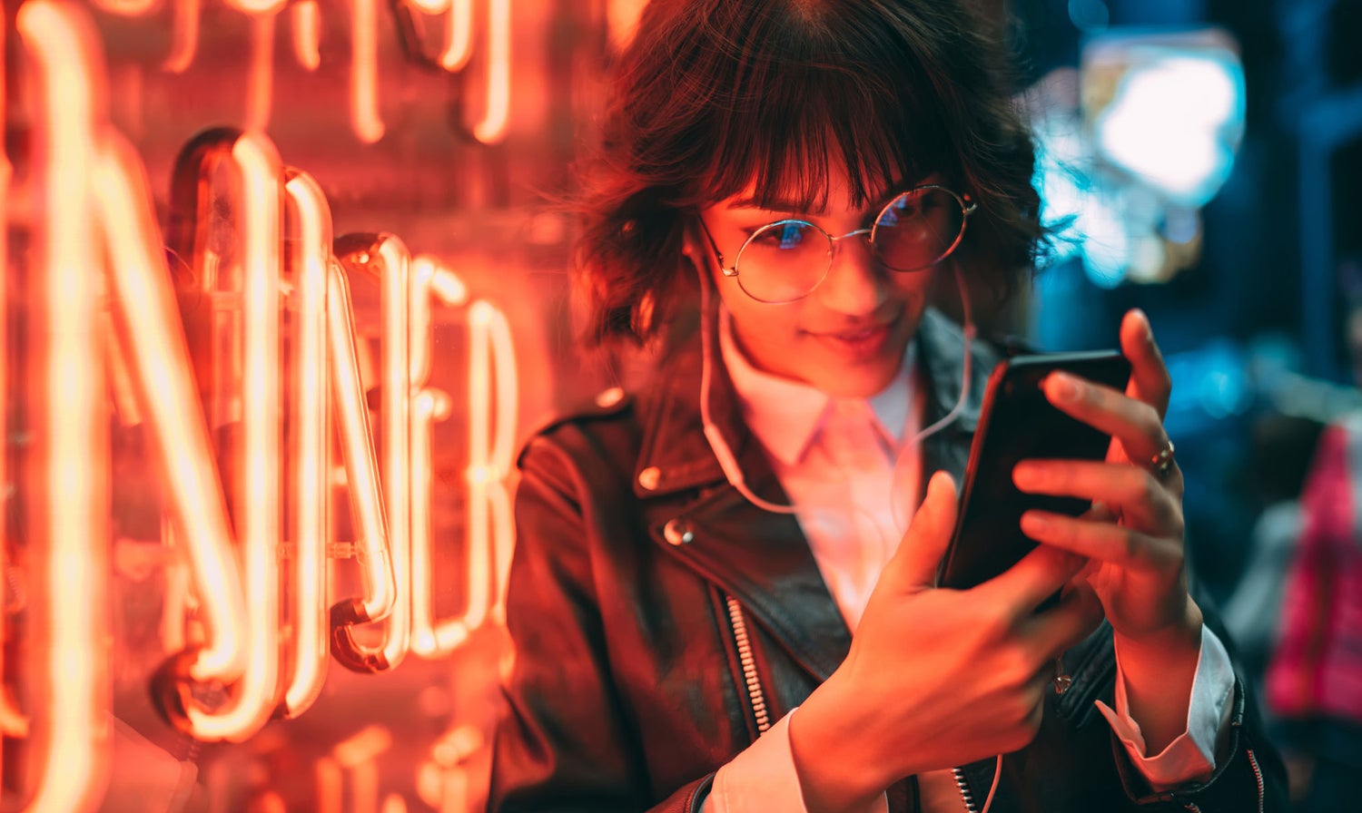 Woman standing in front of a neon sign looking at her phone, with headphones on, wearing a leather jacket.